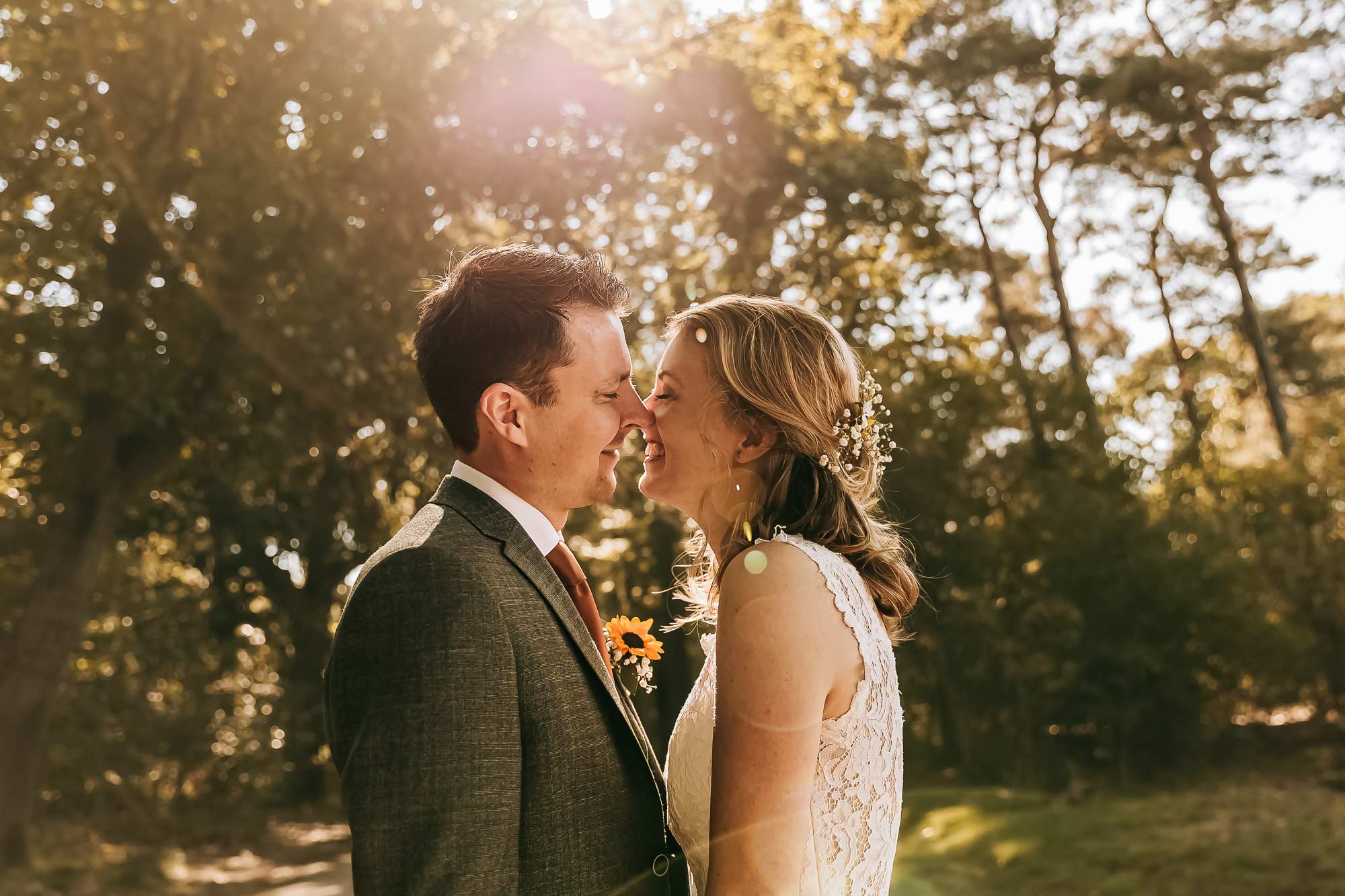 Een mooie foto van een bruidspaar die naar elkaar kijken en elkaar eigenlijk een kus willen geven. De zon schijnt tussen de bomen door waardoor je een lensflare ziet.