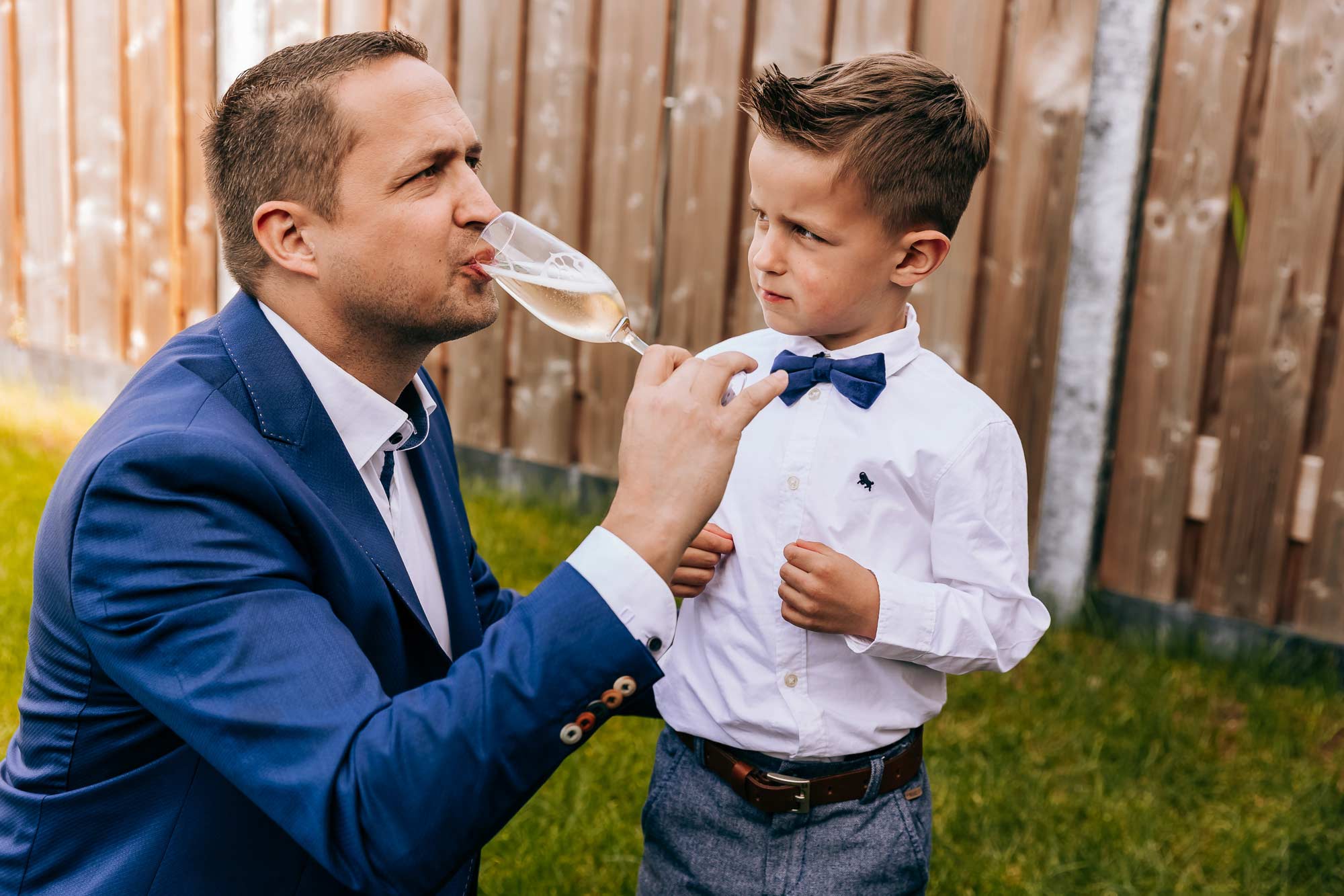 Een kindje op een bruiloft kijkt hoe dat papa champagne uit een glas drinkt, omdat hij zelf eigenlijk ook wel wil proeven.