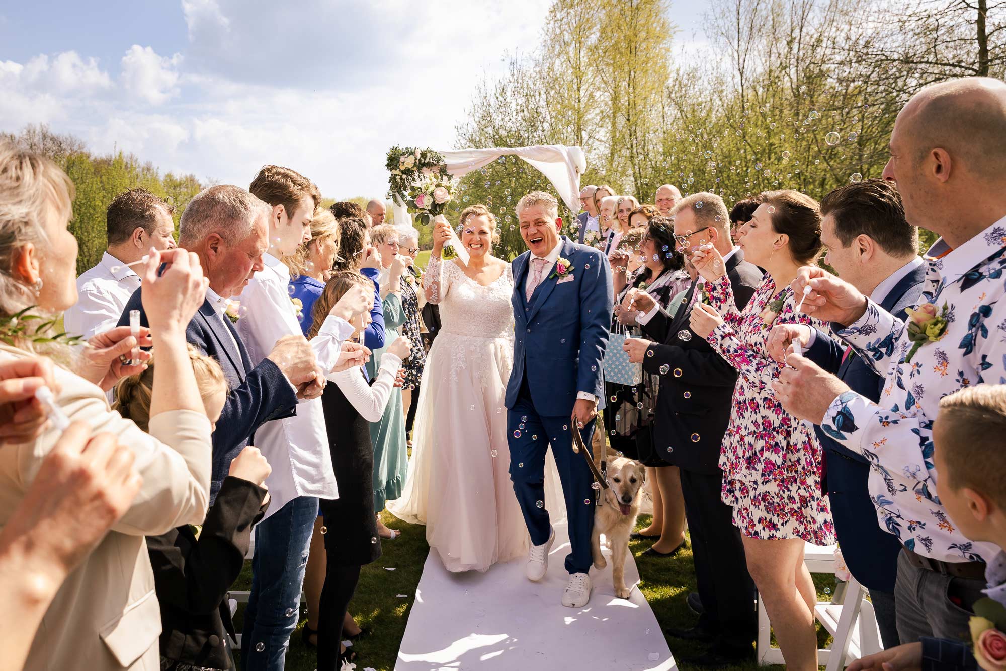 Bruidspaar die tussen de gasten in een haag doorloopt samen met de hond onderweg naar de toost en taart.